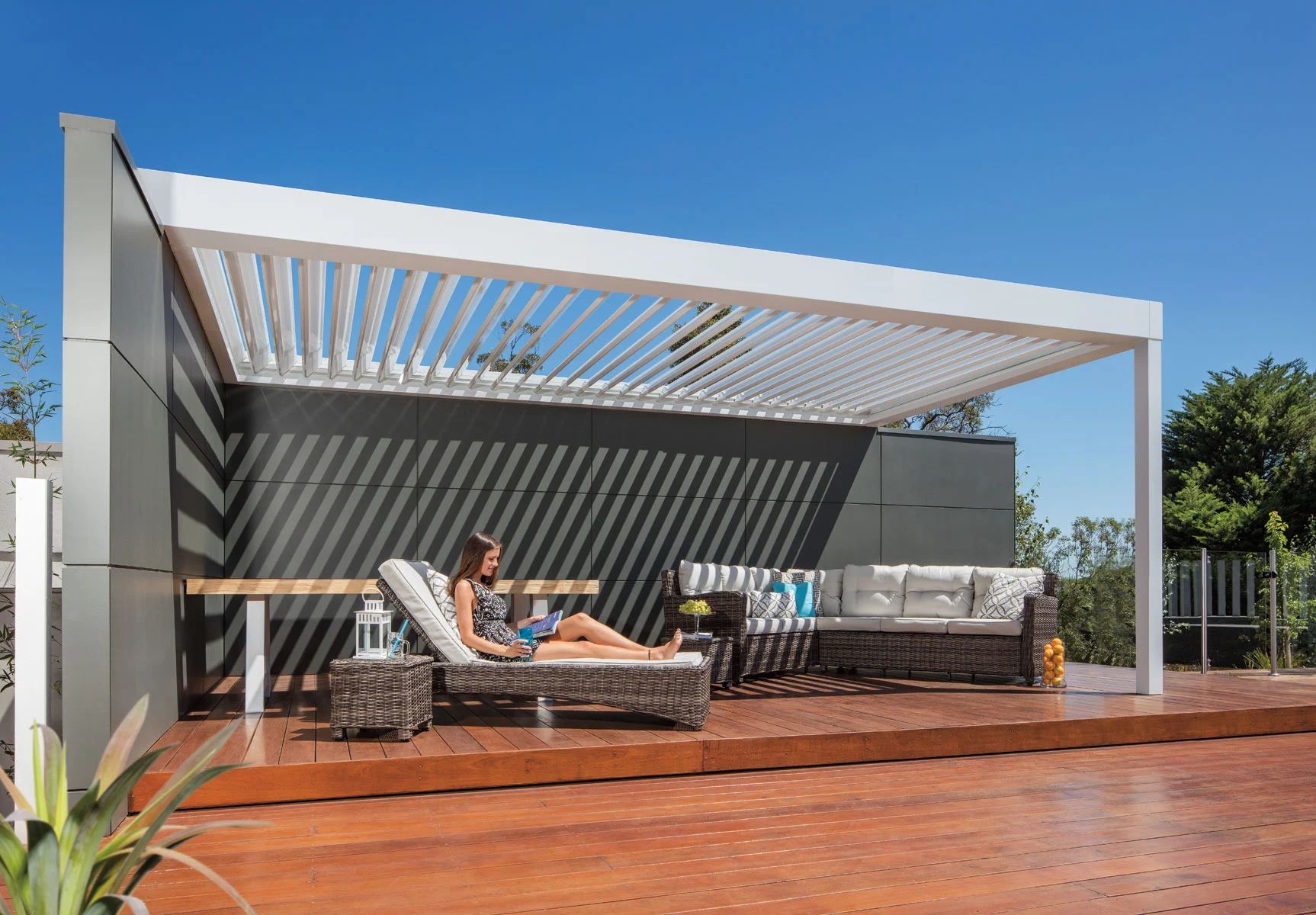 Modern patio with a white pergola, wicker lounge chairs, and sunlit wooden deck under a blue sky