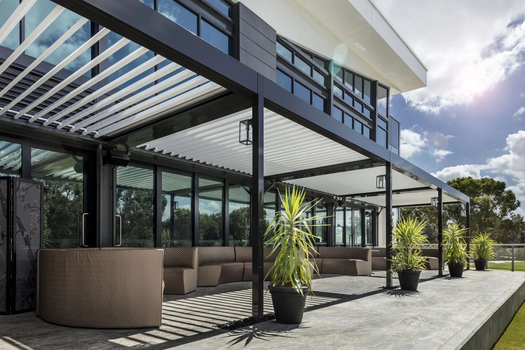 Modern outdoor patio with pergola, cushioned seating, potted plants, and glass walls under a bright sky