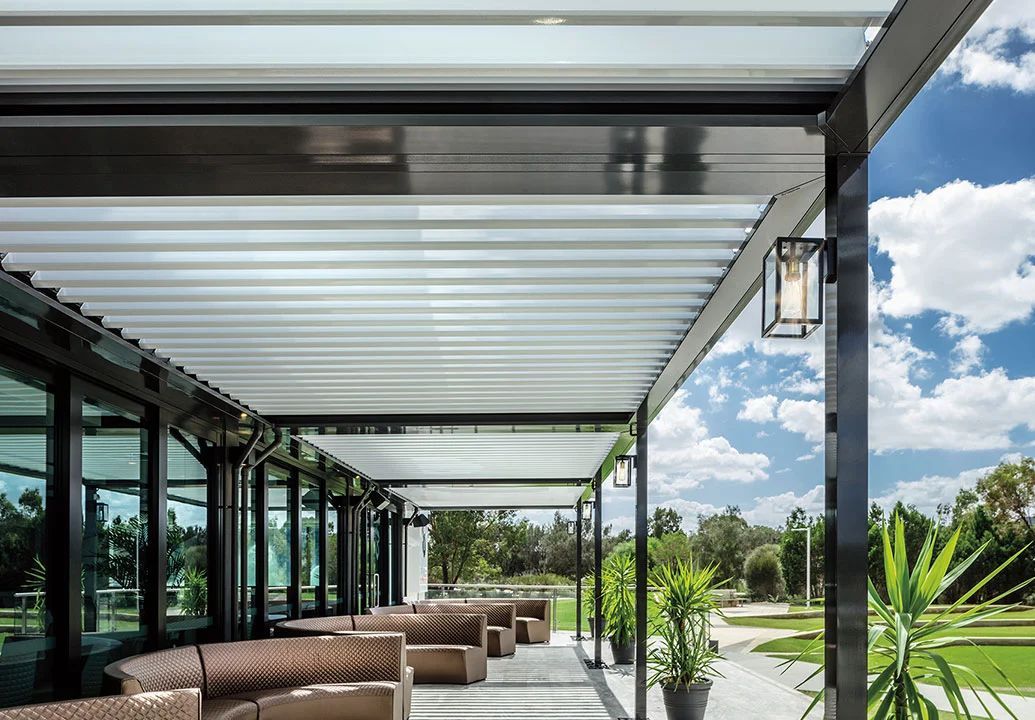 Modern patio with corrugated roof, glass walls, wicker seating, and potted plants under a bright sky