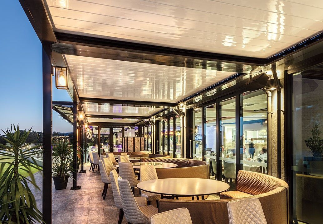 Covered outdoor restaurant seating with round tables, woven chairs, and glass walls at dusk