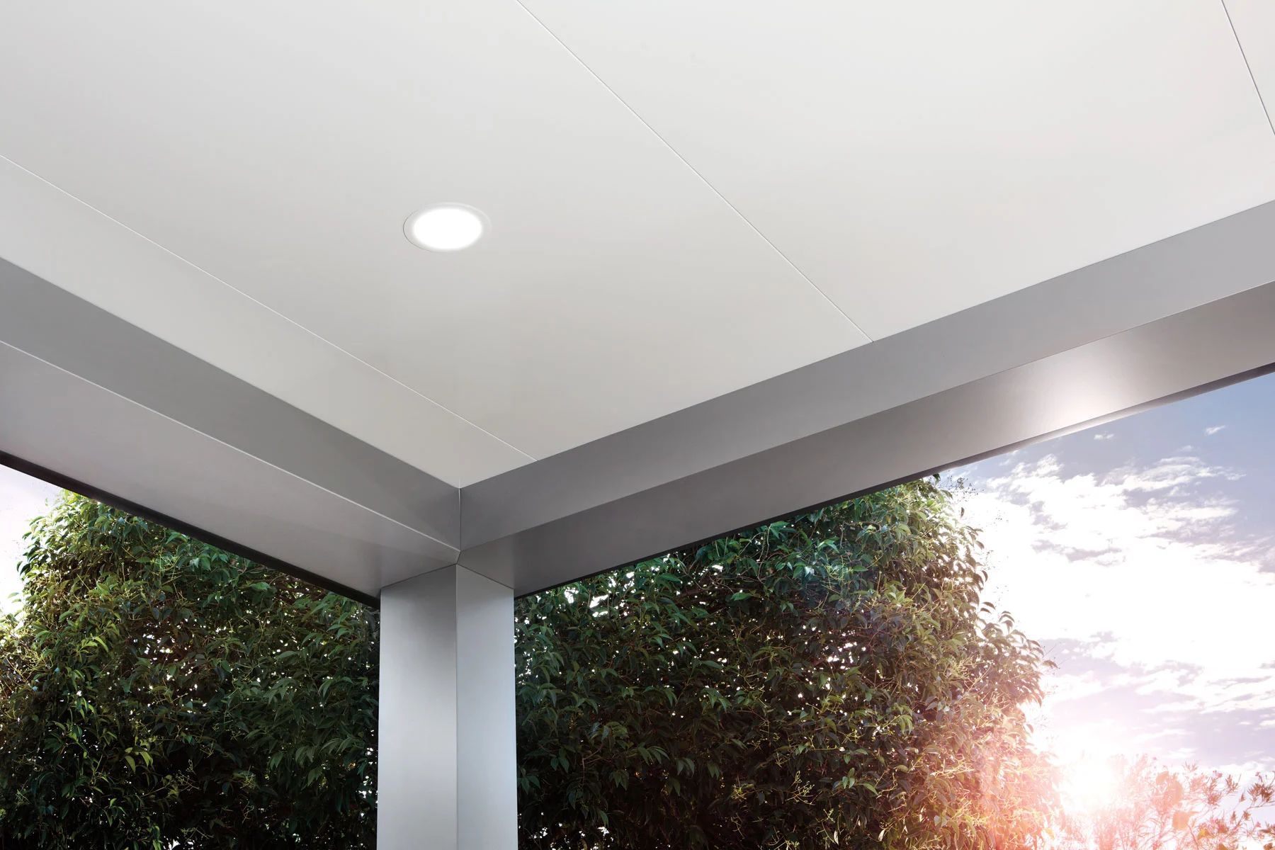 Covered patio ceiling with recessed light, framed by gray beams and trees at sunset