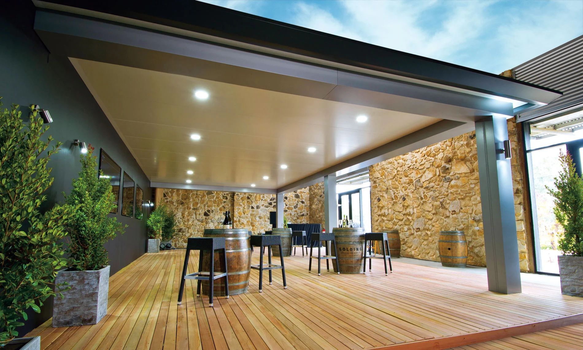 Covered wooden patio with bar stools and tables, lit by recessed lights, beside a stone wall and greenery.