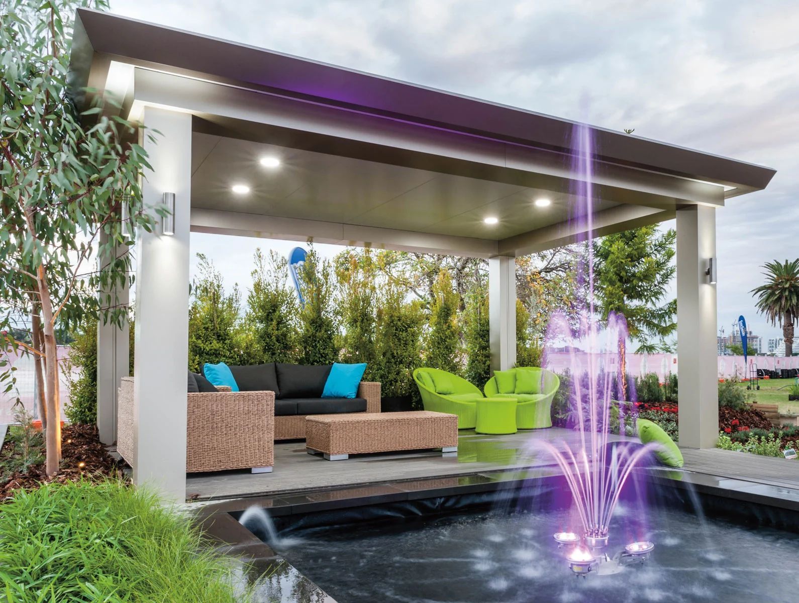 Modern backyard patio with wicker seating, green chairs, and a lit fountain beside a covered pergola