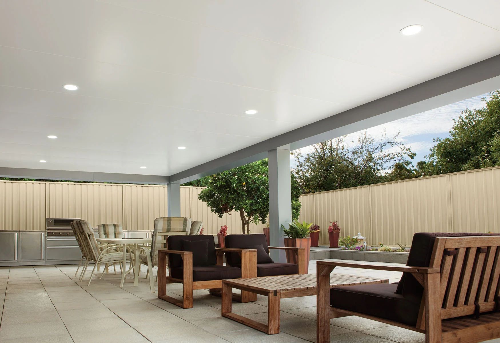 Covered outdoor patio with wooden seating, dining table, and potted plants under a white ceiling