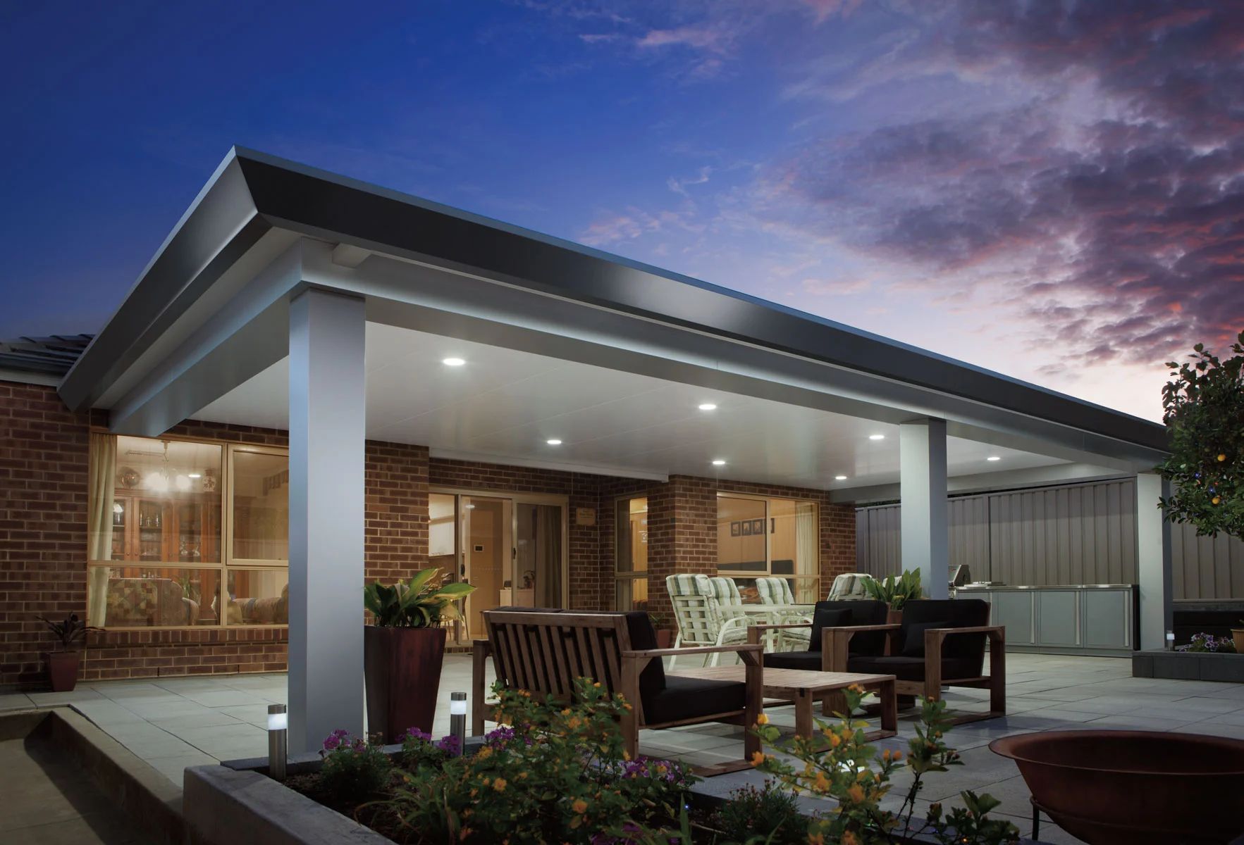 Modern patio with outdoor dining set under a lit roof at dusk beside a brick house