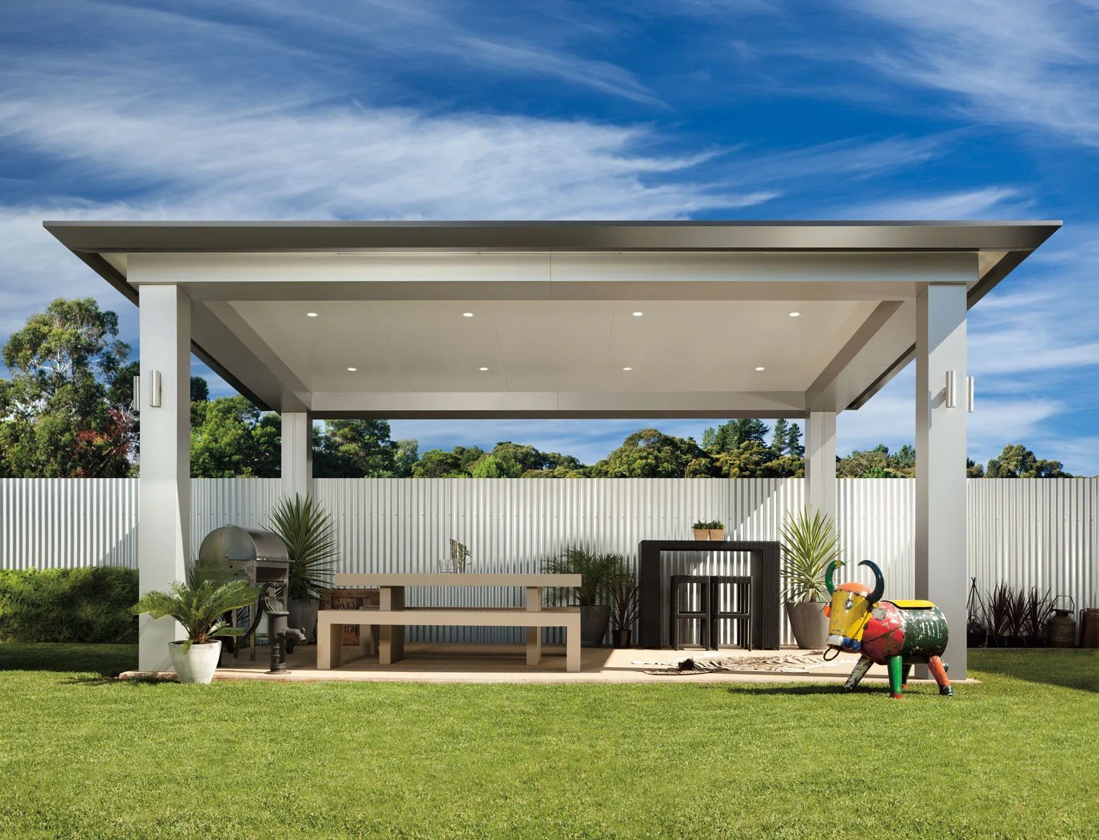 Modern backyard patio with a covered seating area, grill, and green lawn under a blue sky