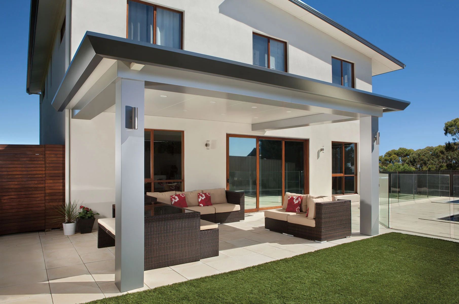 Modern two-story house with a covered patio, outdoor seating, and green lawn in bright sunlight