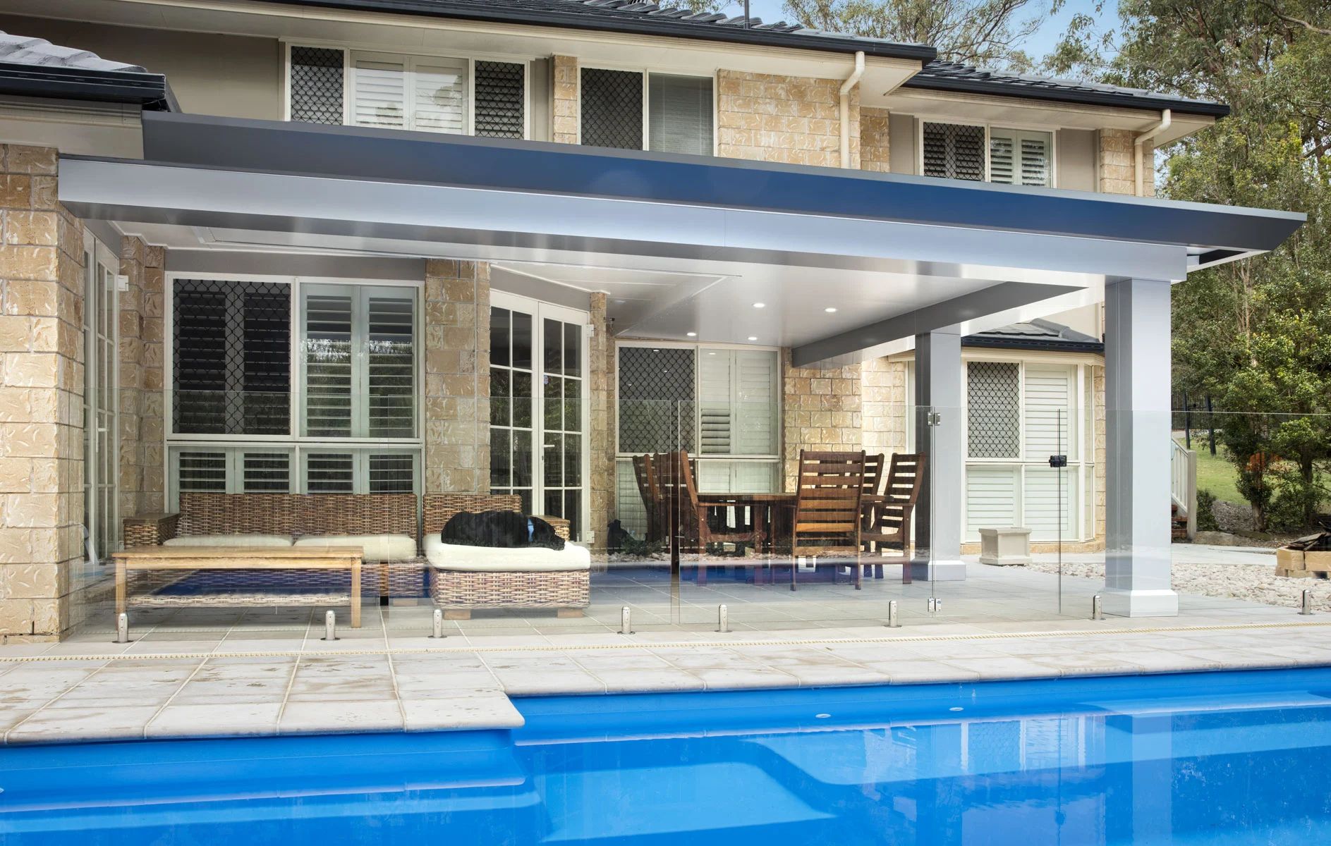 Modern patio with seating under a covered awning beside a blue swimming pool and house exterior