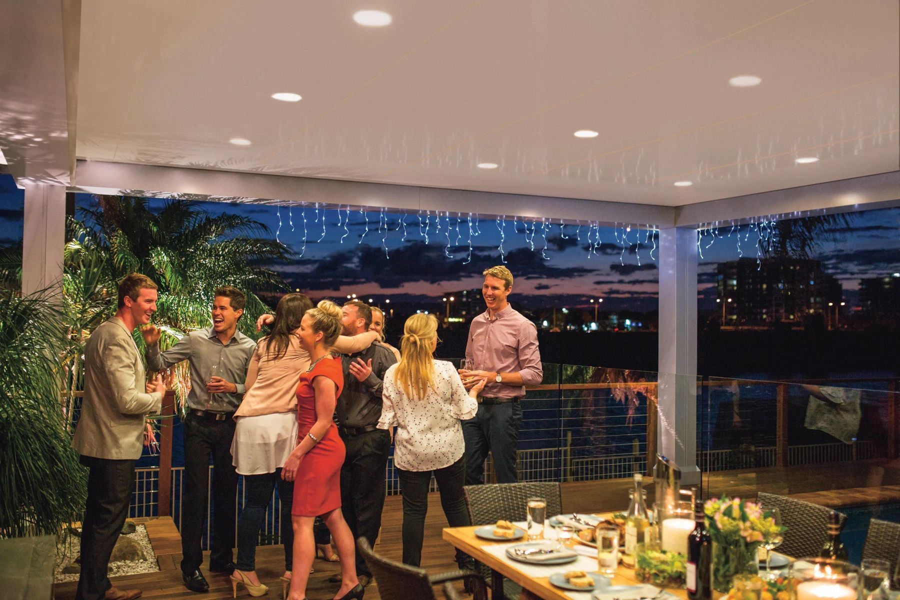 Guests mingling on a rooftop terrace at sunset with city lights and a set dining table