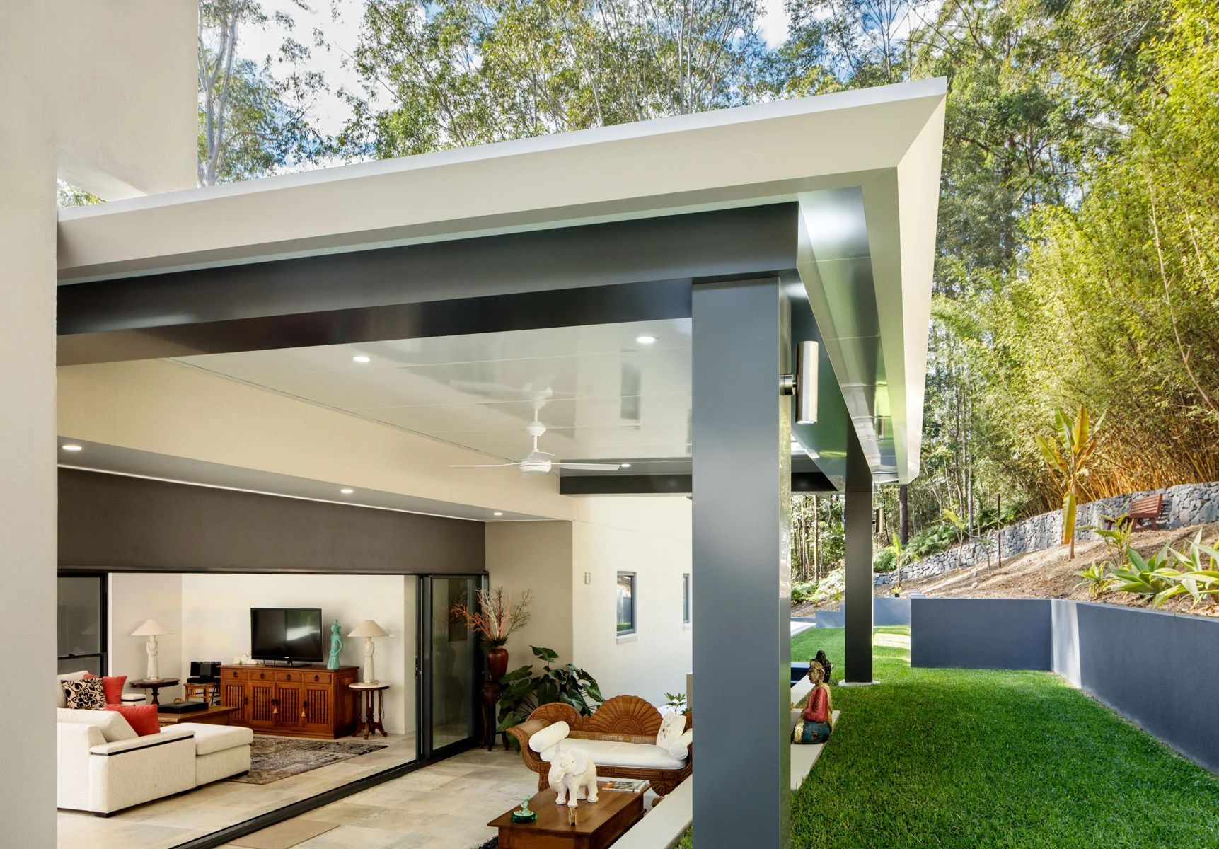 Modern house patio with open sliding doors, white roof, and green lawn beside a hillside.