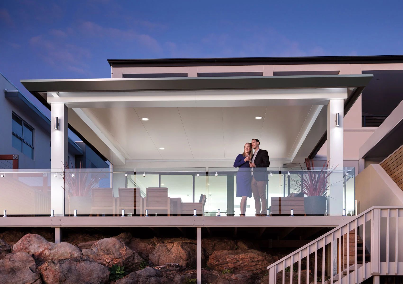 Modern house patio with glass railing; two people stand on the balcony at dusk.
