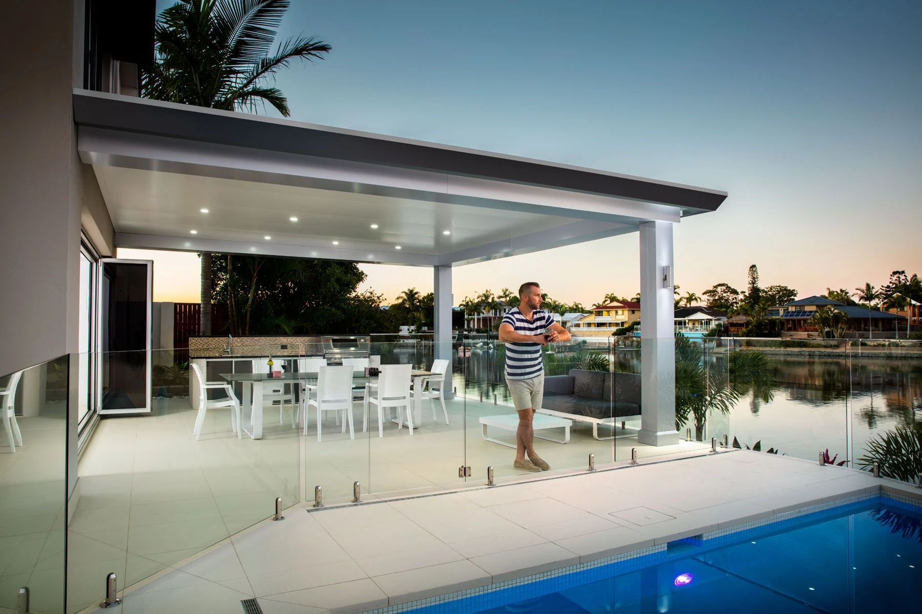 Modern patio with white furniture, pool, and one person standing by a waterfront at sunset
