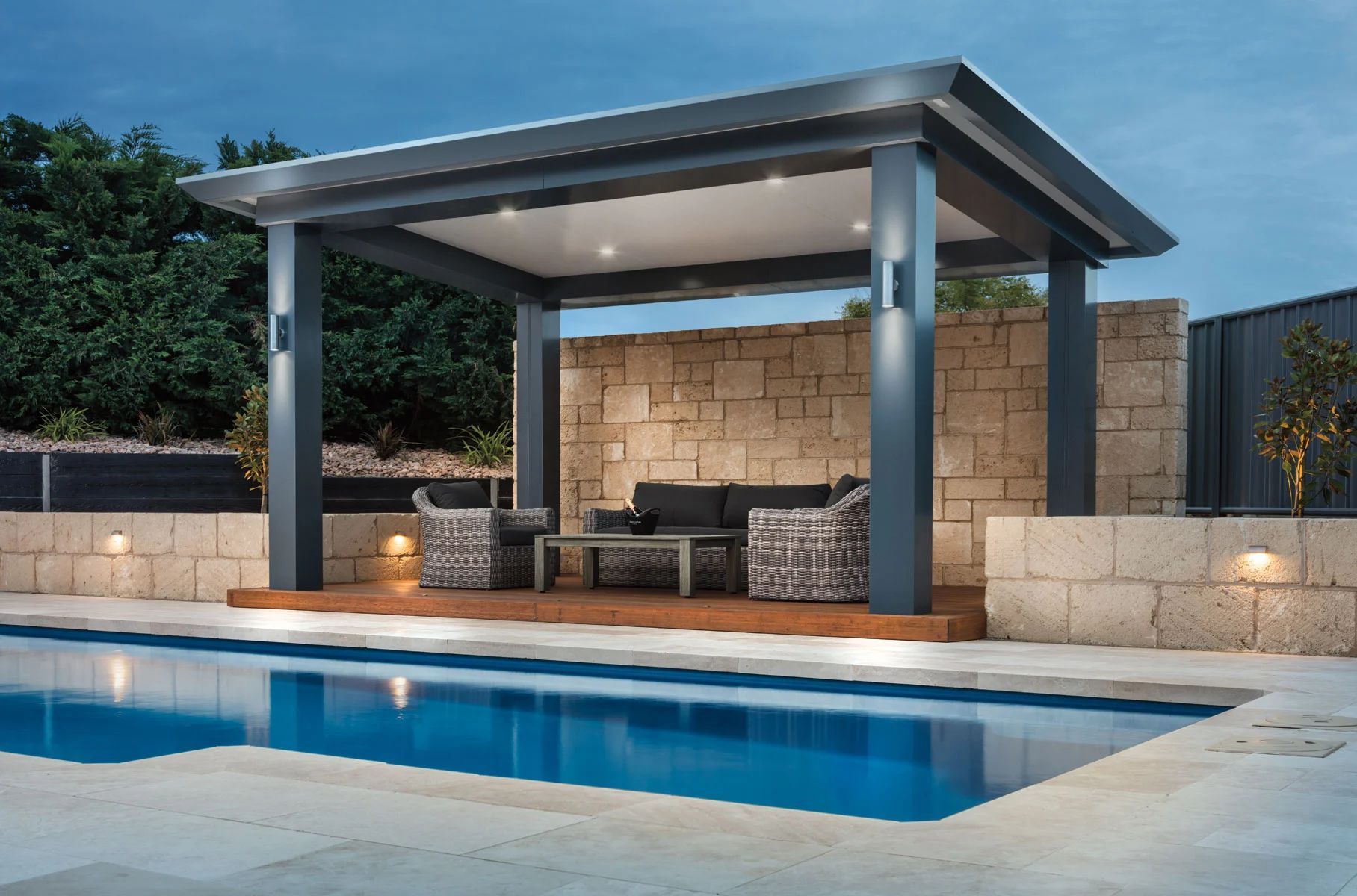 Modern poolside cabana with stone backdrop, outdoor seating, and blue swimming pool at dusk