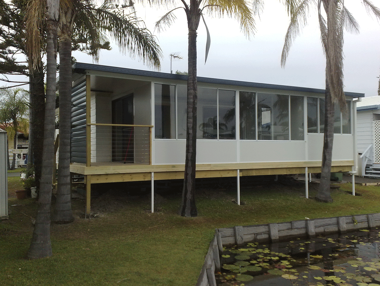 Small white stilt house with large windows and palm trees beside a pond.