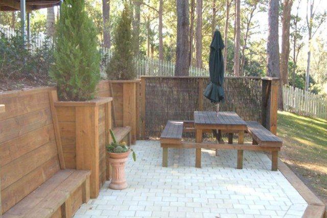 Back patio with wooden bench, picnic table, closed umbrella, potted plant, and fence under trees