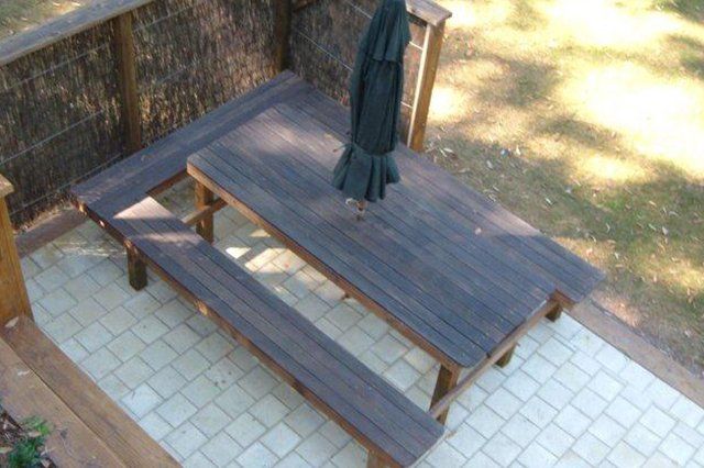 Wooden picnic table with a closed umbrella on a sunlit patio beside a stone wall
