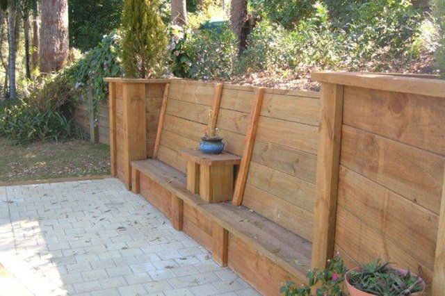 Wooden outdoor bench along a paved patio beside a garden, with a small blue planter on a central post.