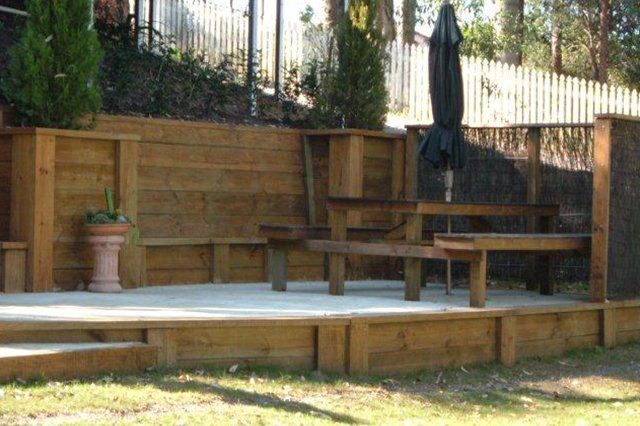 Wooden deck with benches, potted plants, and a closed patio umbrella in a fenced backyard.