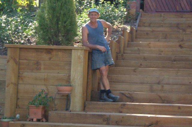 Person in blue tank top and shorts leaning on a wooden fence beside outdoor stairs.