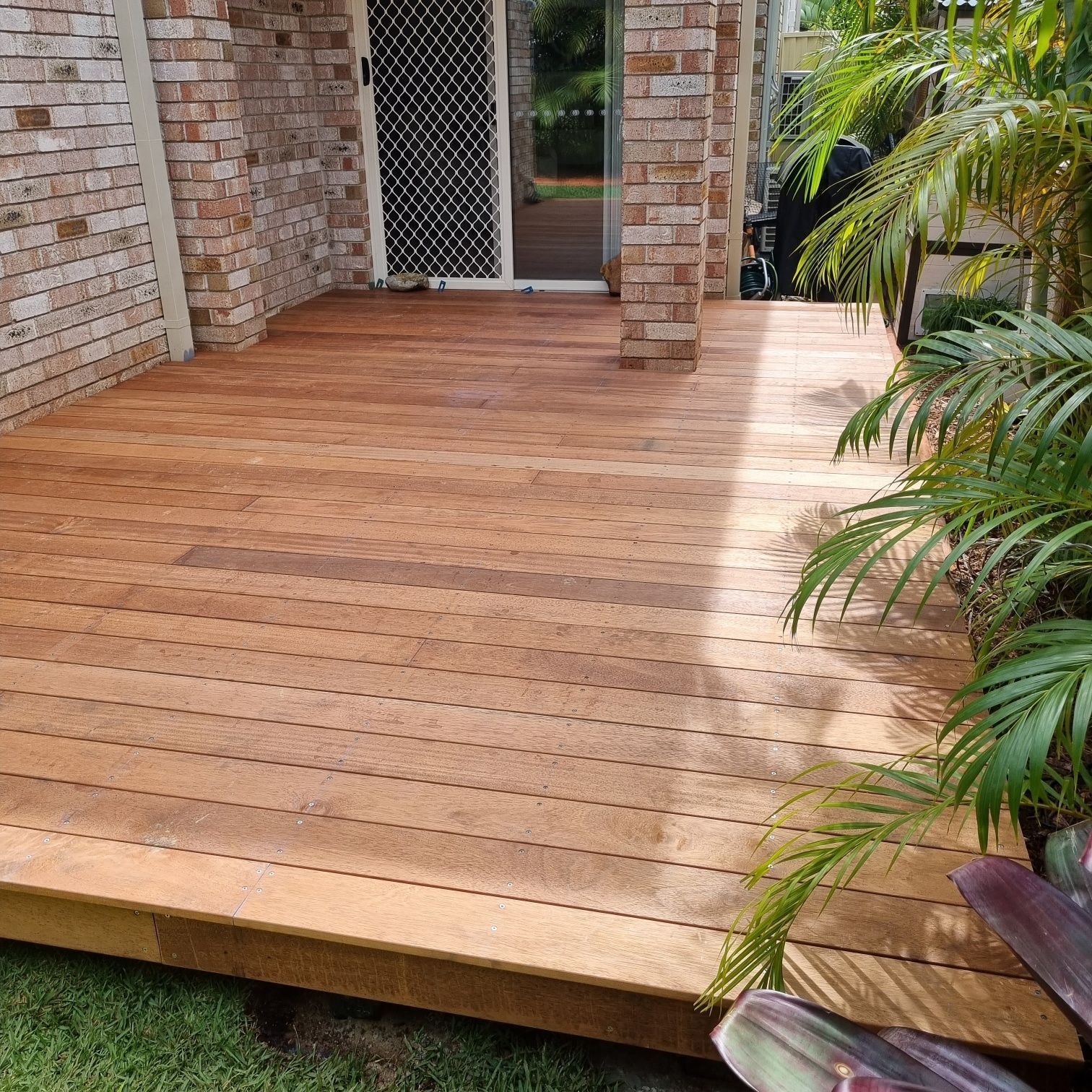 Wooden deck beside a brick house with a glass door and tropical plants nearby
