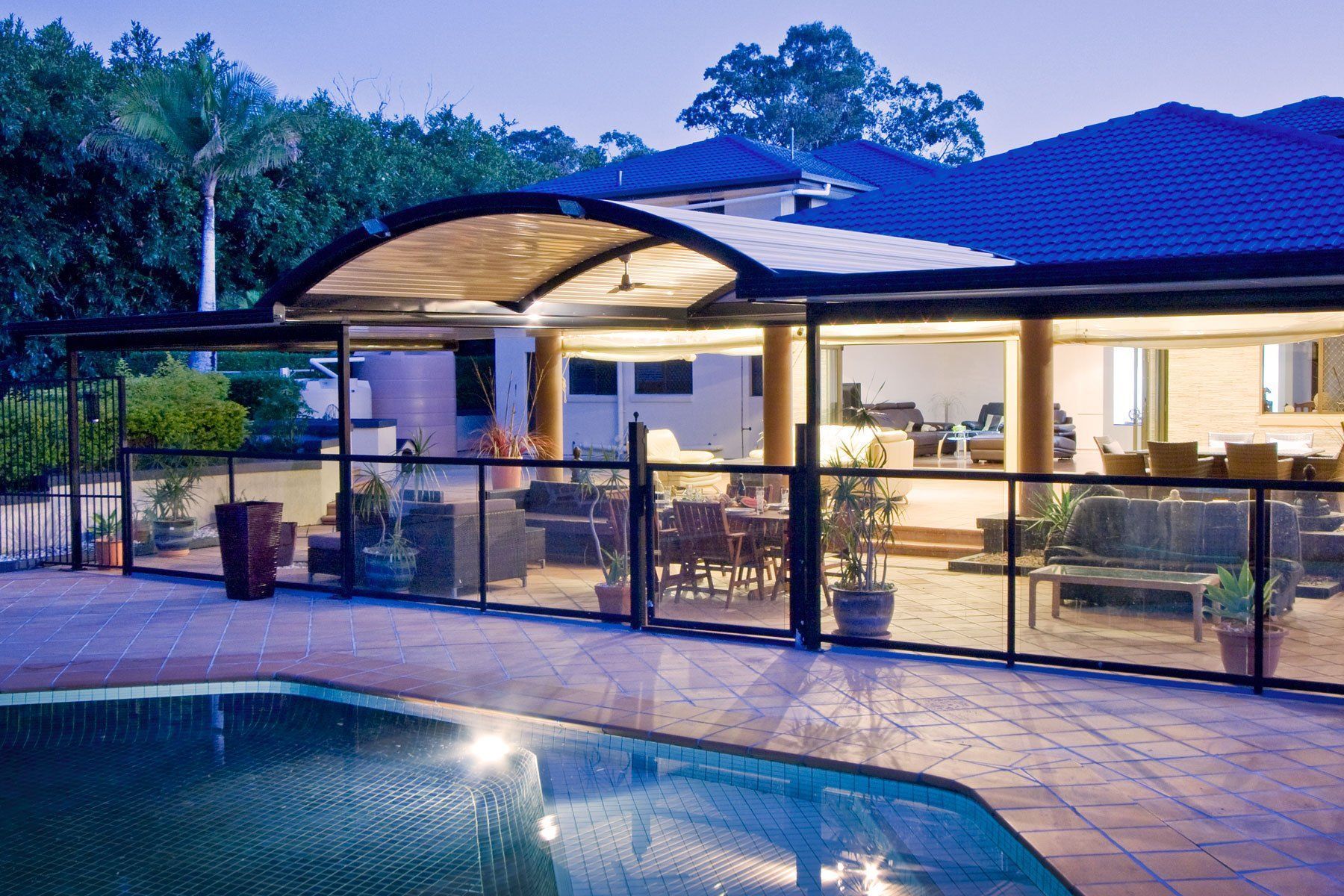 Backyard pool beside a lit patio and modern house at dusk