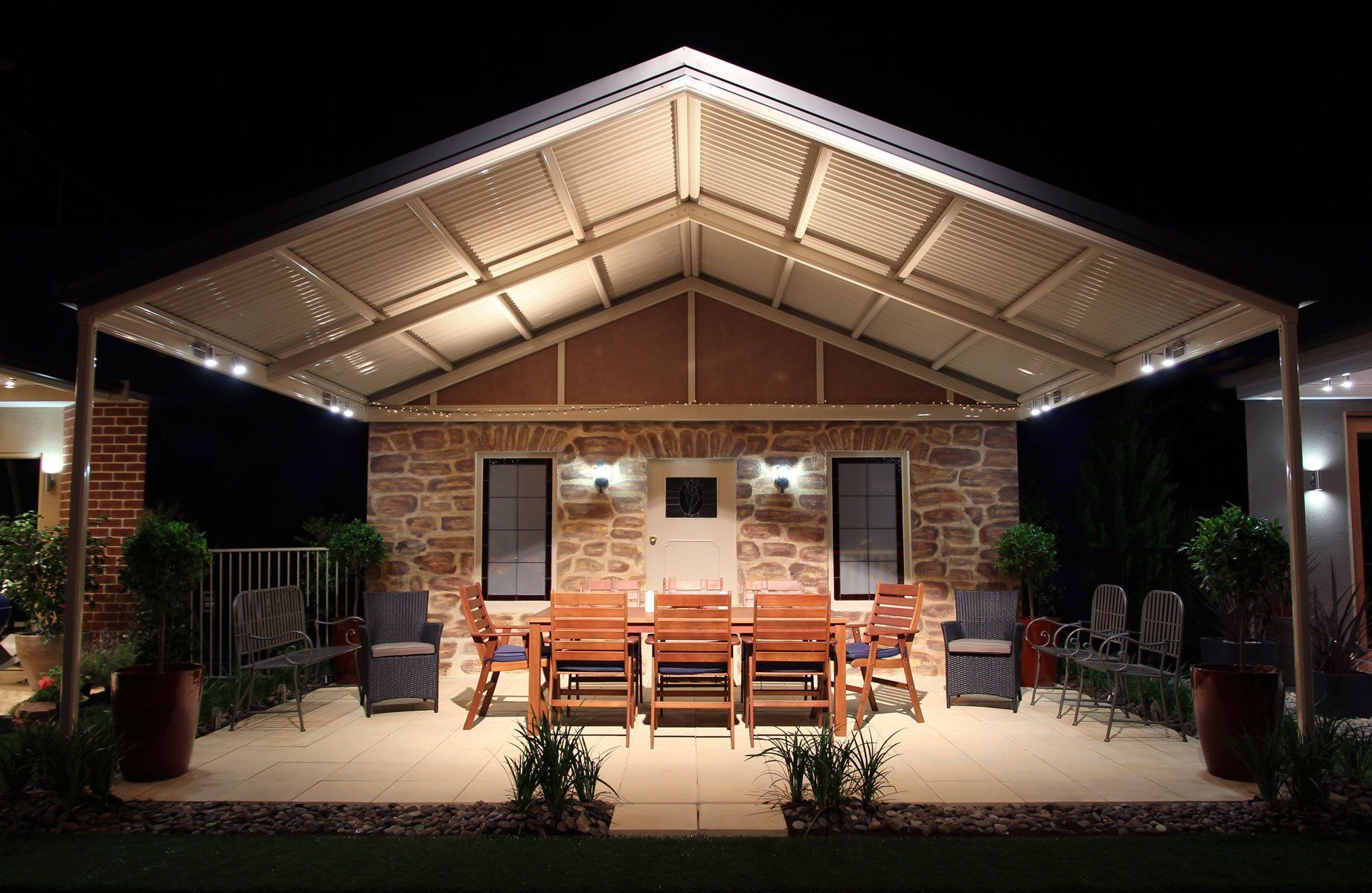 Brick patio home with lit covered porch, wooden chairs, and potted plants at night