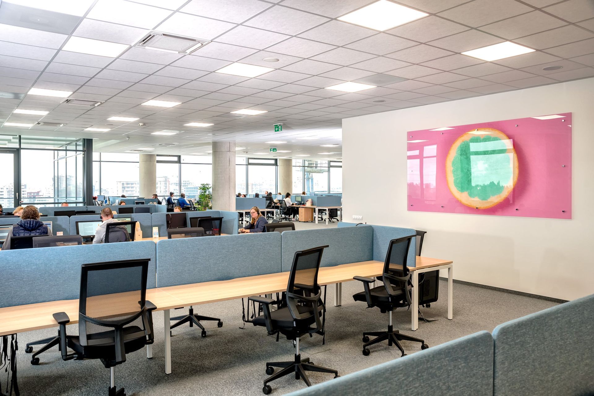 Modern office lounge with blue workstations, black chairs, and a pink wall art panel