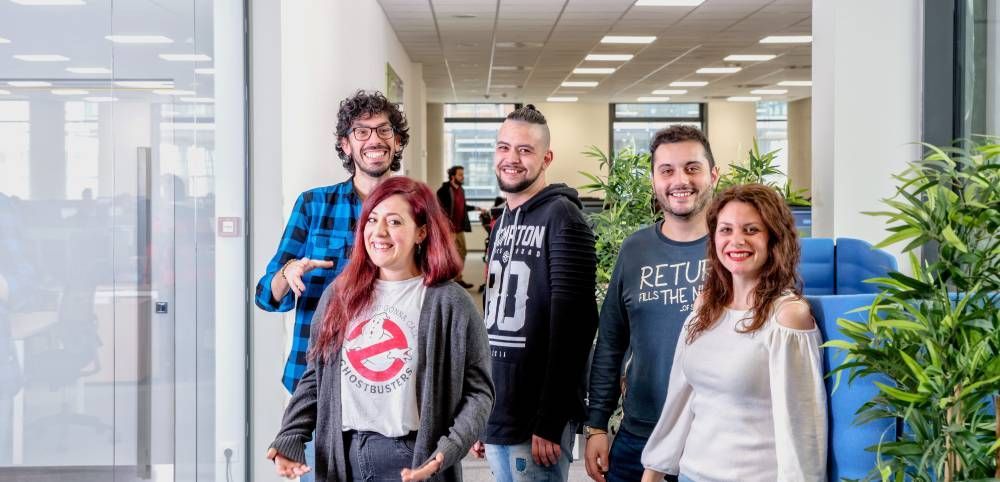 Five coworkers standing in a bright office hallway, smiling at the camera