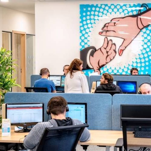 Open office with people working at computers beneath a colorful mural of hands holding a heart-shaped chain