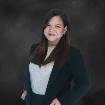 Woman in a black blazer and white top, standing against a dark studio backdrop and smiling softly