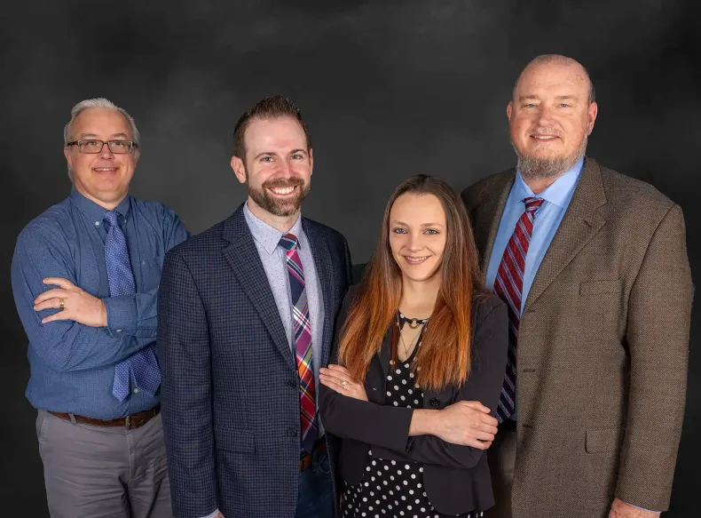 Four people posing formally against a dark gray backdrop, smiling with arms crossed.