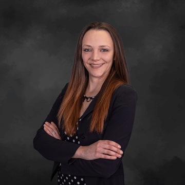 Woman with long brown hair in a black blazer, smiling with arms crossed against a dark gray studio backdrop.