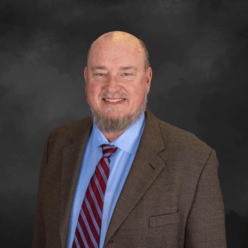 Smiling bald man in a brown blazer, blue shirt, and red striped tie against a dark background