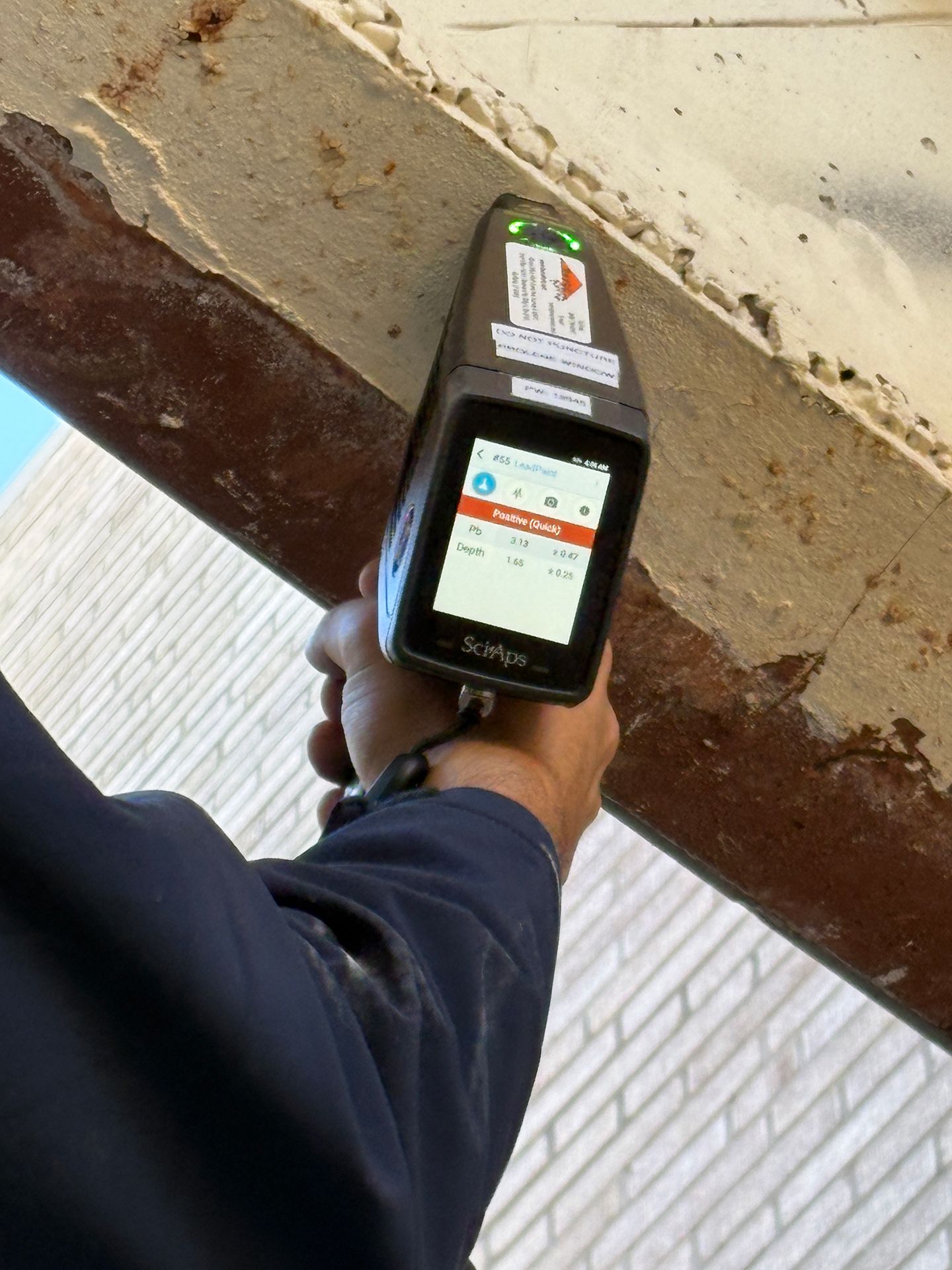 Handheld device scanning a metal beam under a structure, with a warning label visible on the beam
