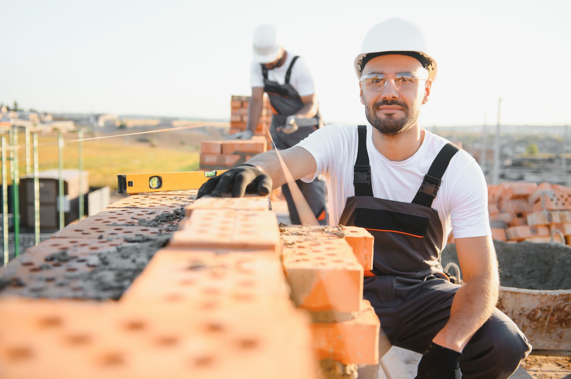 Construction worker smiling, laying bricks on a building site. Another worker in the background.