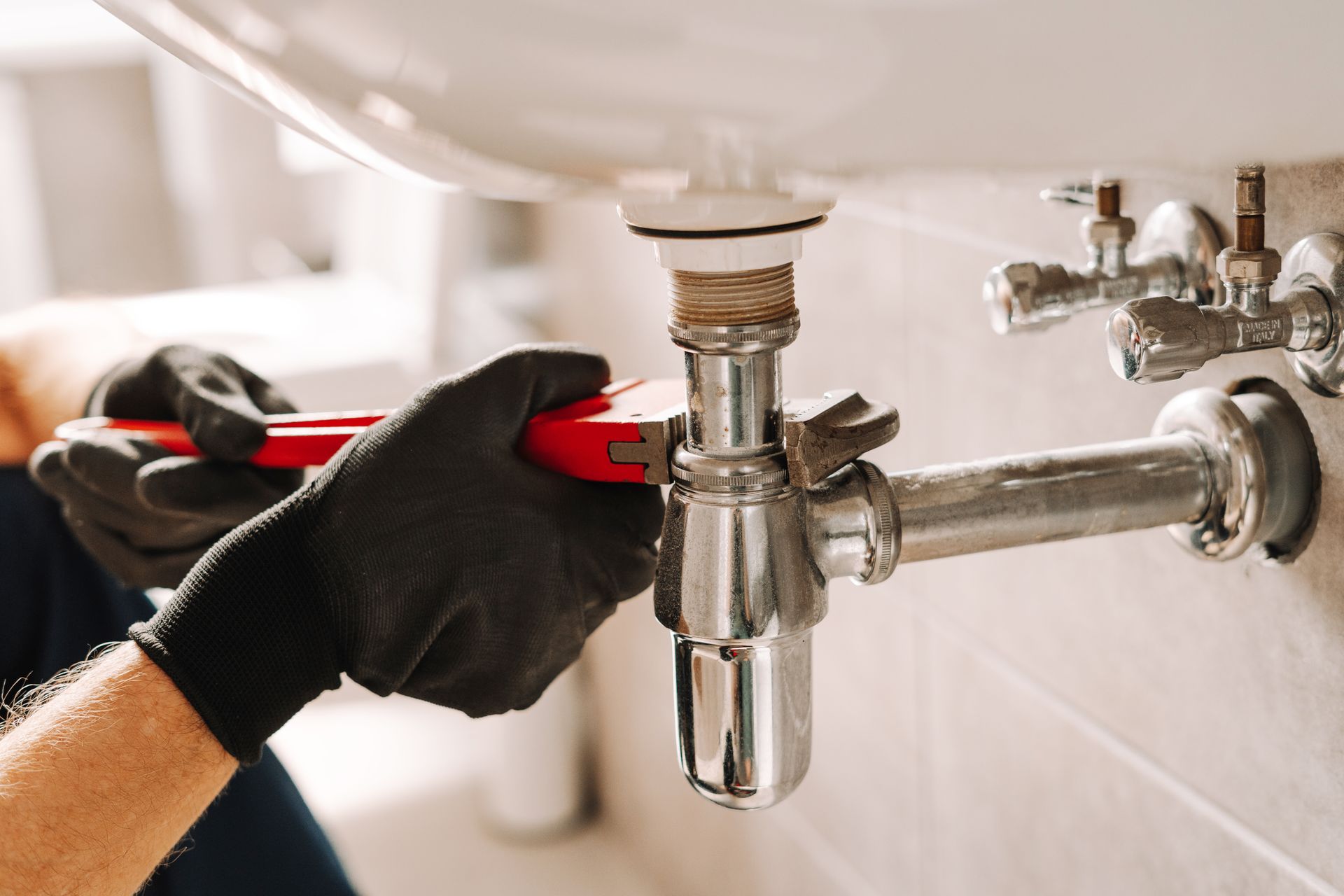 Plumber wearing black gloves uses a wrench to tighten a pipe under a white sink in a bathroom.