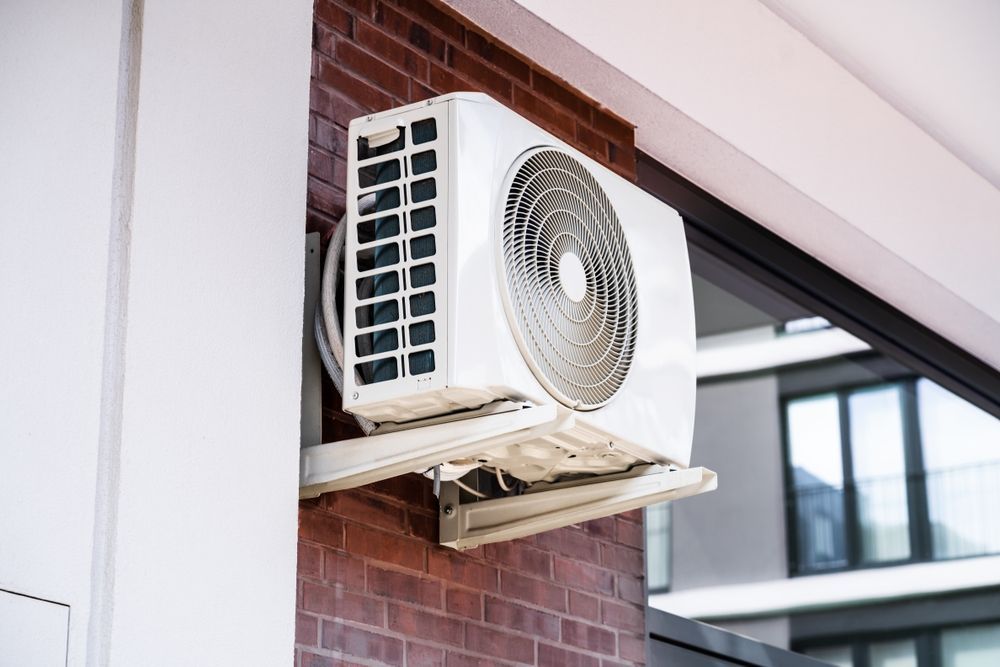A white air conditioning condenser unit mounted on a red brick exterior wall next to a glass window.