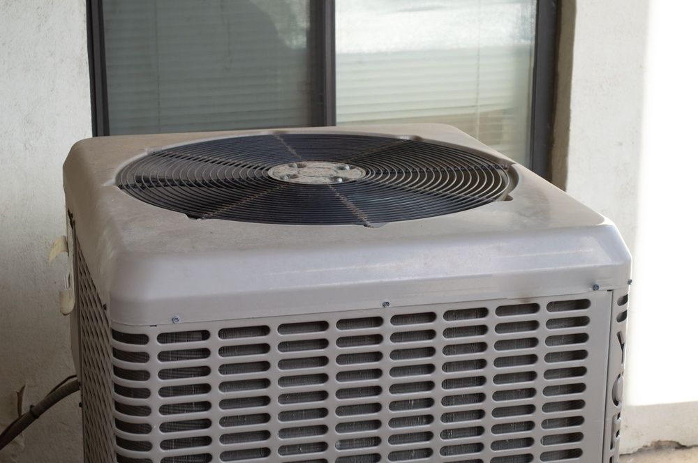 Outdoor air conditioning unit with a black fan grate sitting on a patio next to a glass sliding door.