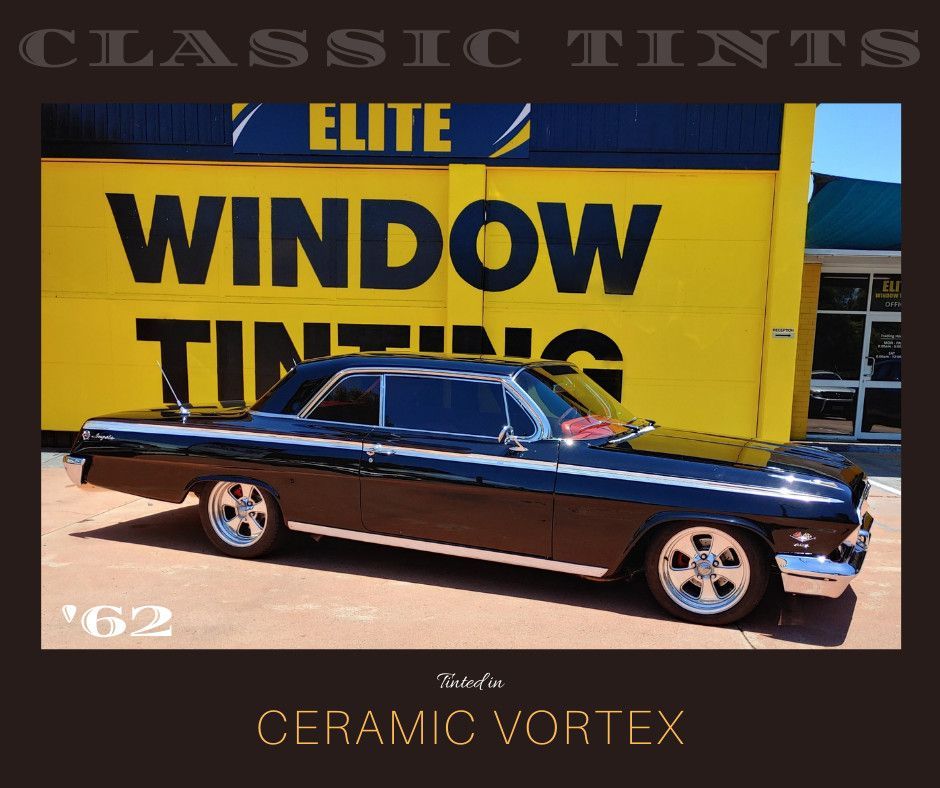 A black 1962 Chevrolet parked in front of a yellow Elite Window Tinting shop wall under 