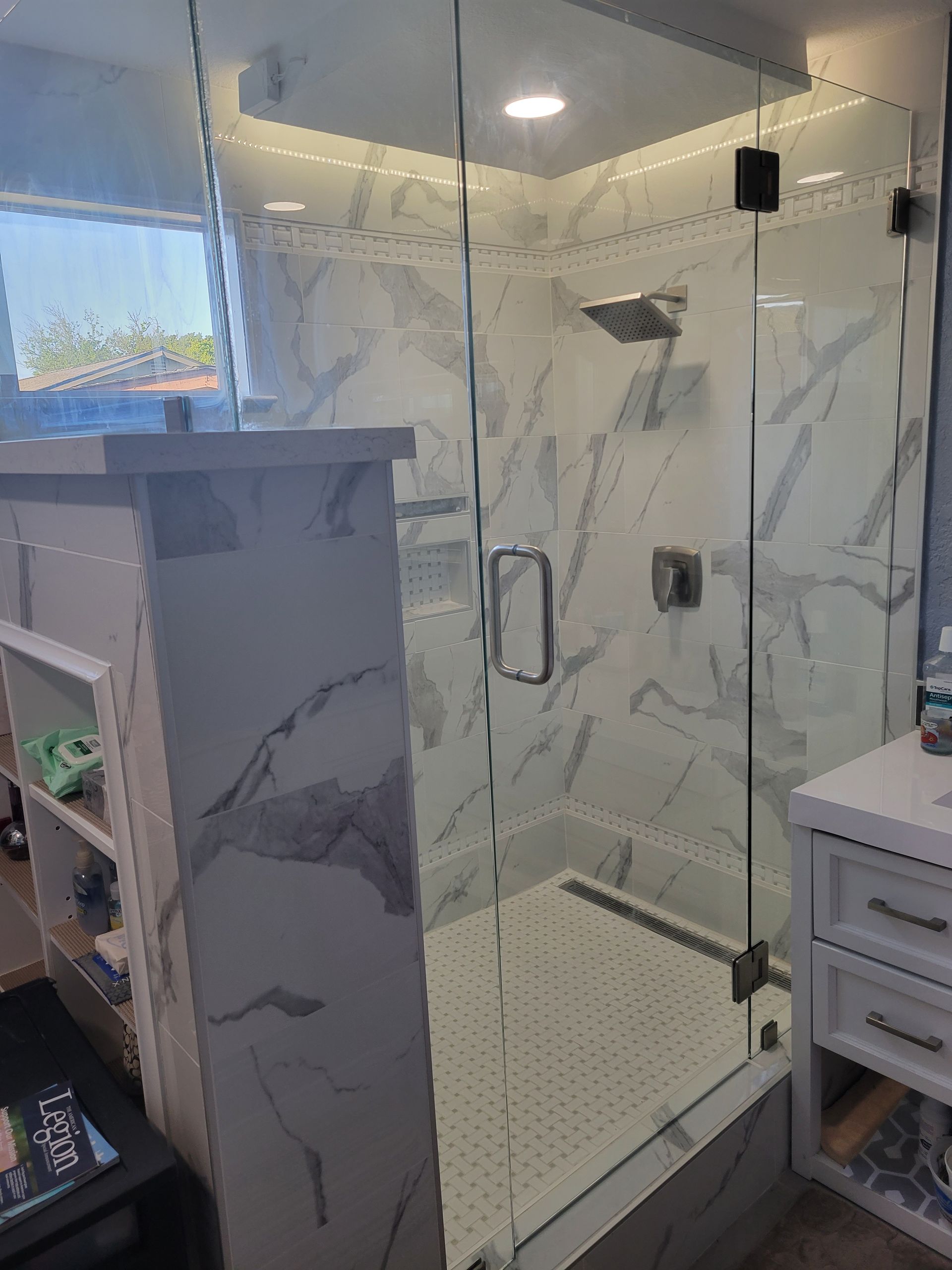 Glass shower enclosure in a marble-tiled bathroom with vanity and mirror lights, viewed from the bedroom.