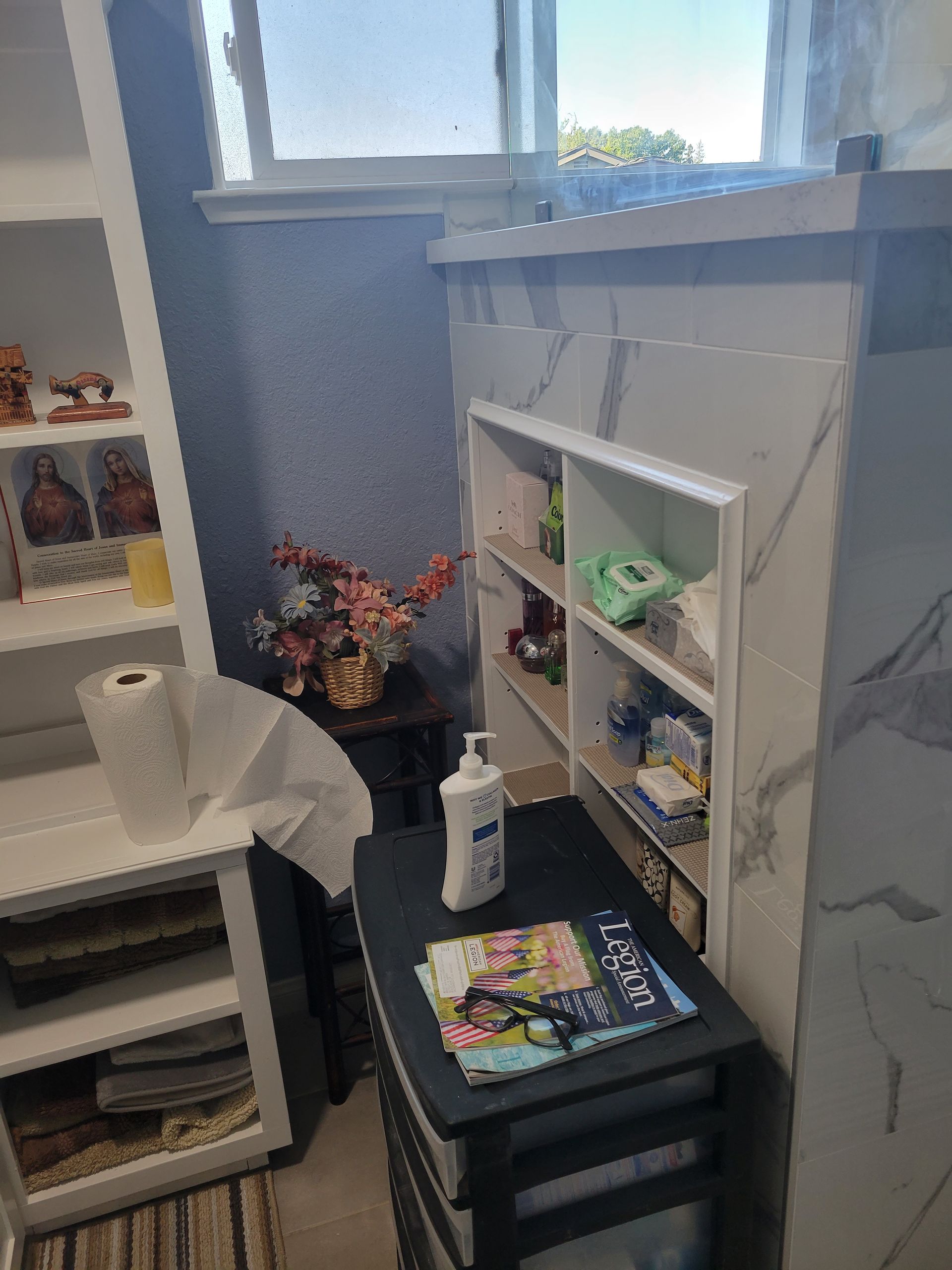 Small room corner with white shelving, black drawers, books, flowers, and a window above a fireplace.
