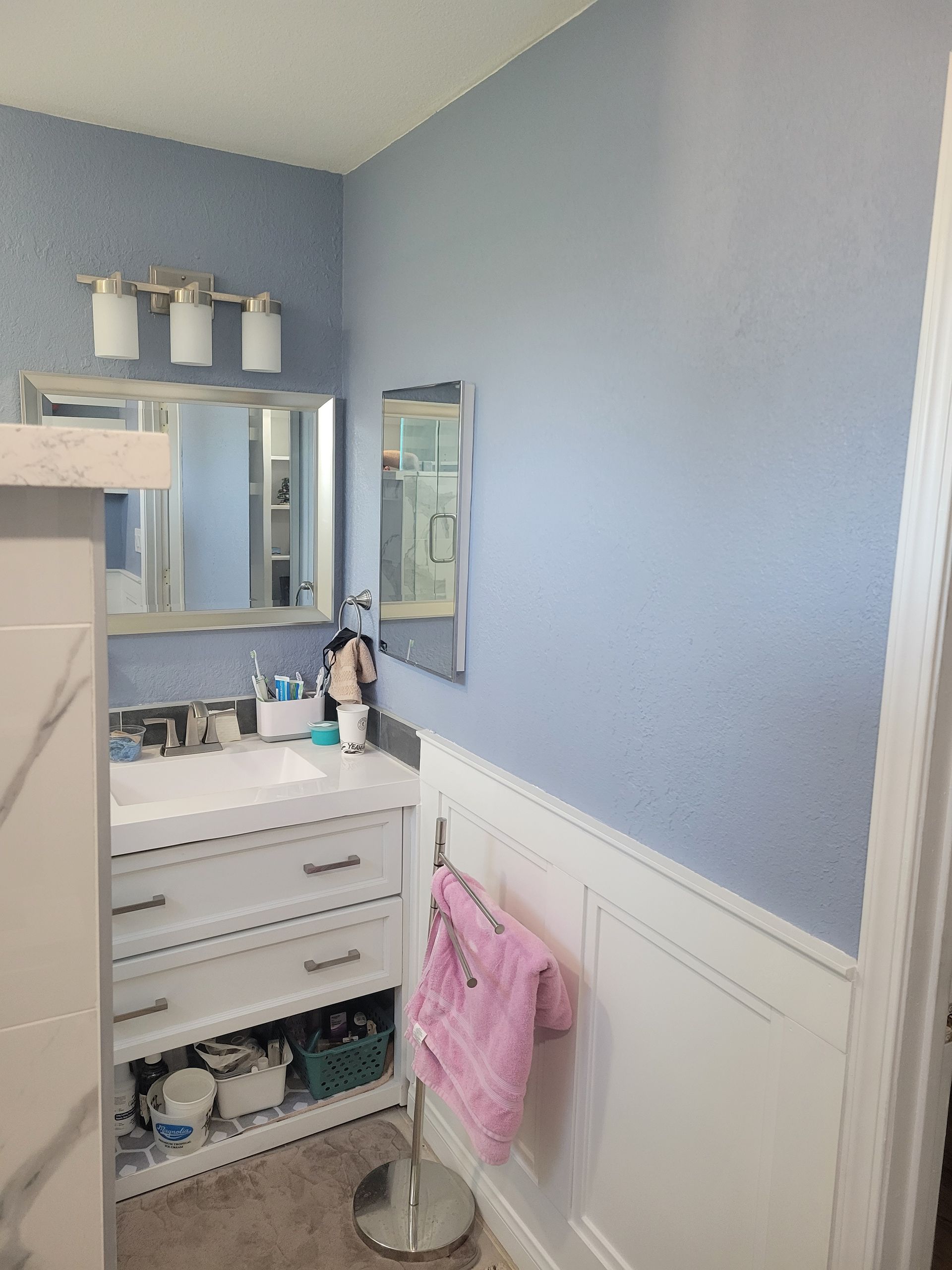 Small blue-and-white bathroom with vanity, mirror, wall paneling, and pink towel on a stand