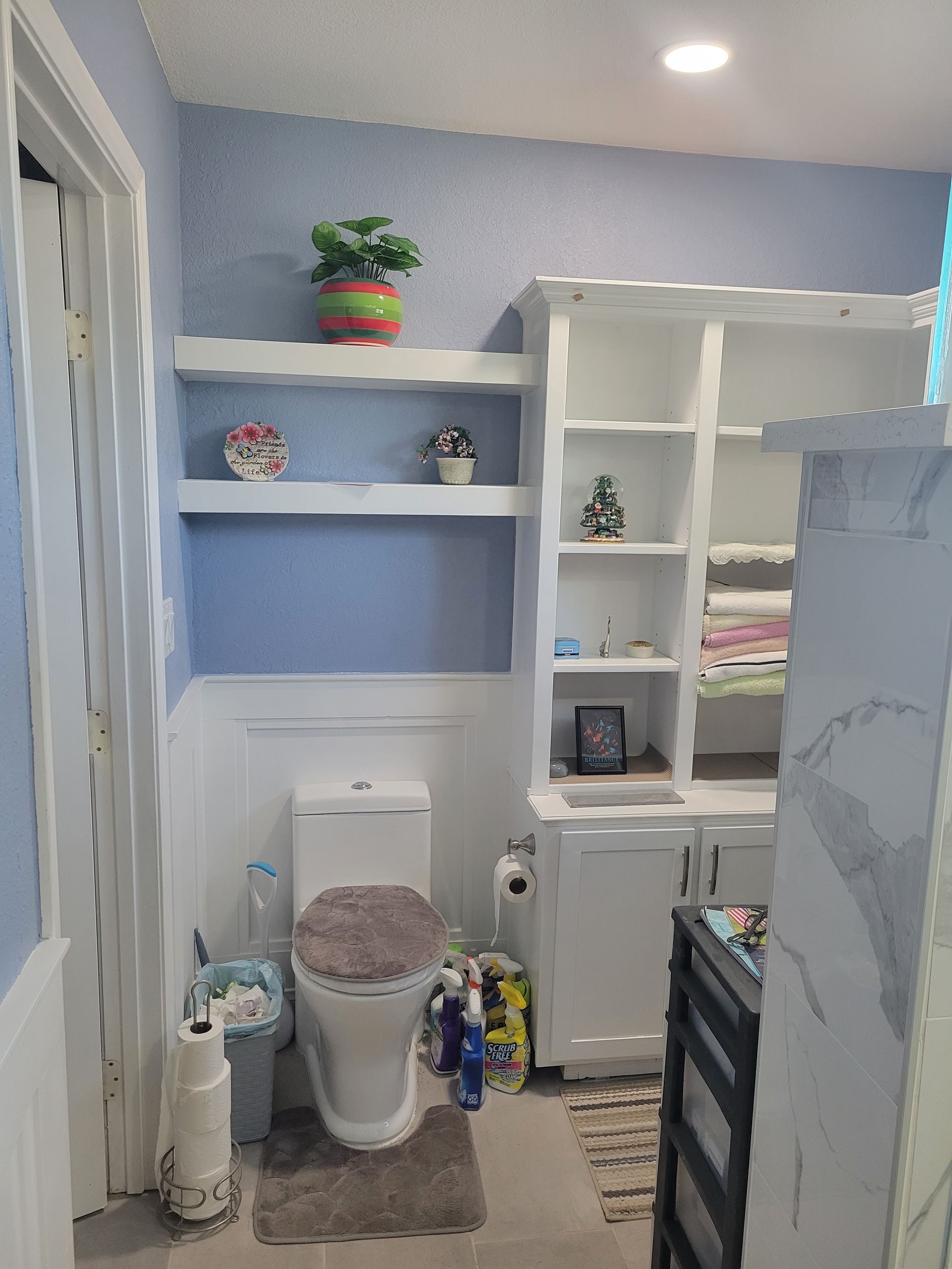 Small blue bathroom with white built-in shelves, toilet, and a marble-look shower wall.