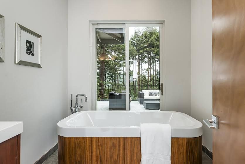 Modern bathroom with a wood-accent tub, open door, and patio view of greenery beyond