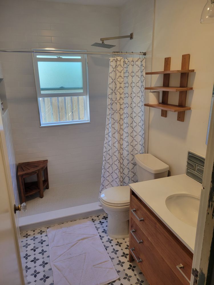 Small bathroom with window, shower curtain, toilet, sink vanity, wall shelves, and patterned floor tile.