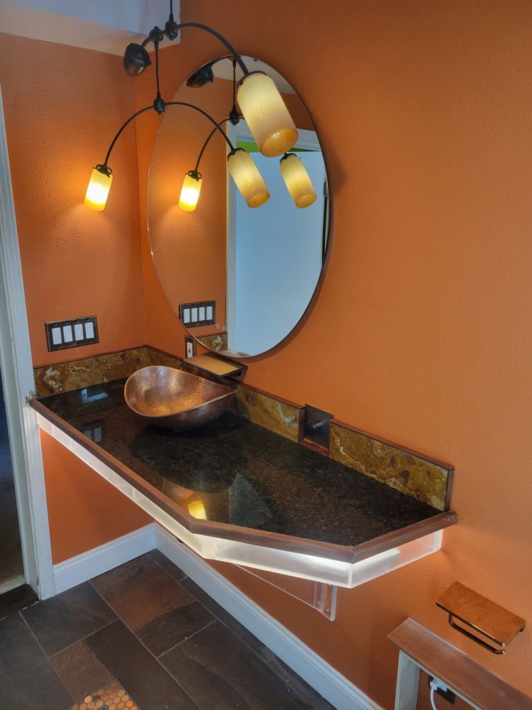 Small bathroom vanity with oval mirror, black granite countertop, and wall sconces against orange walls
