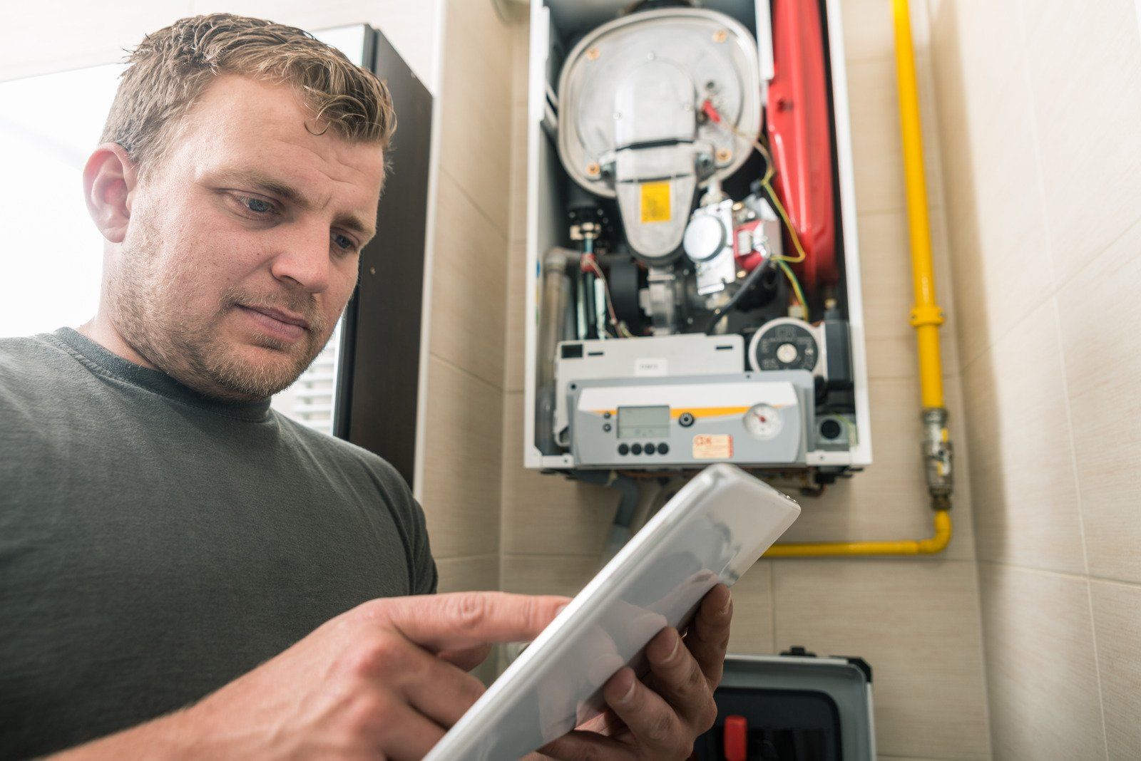 Man looking at tablet, inspecting a gas boiler in a utility room.