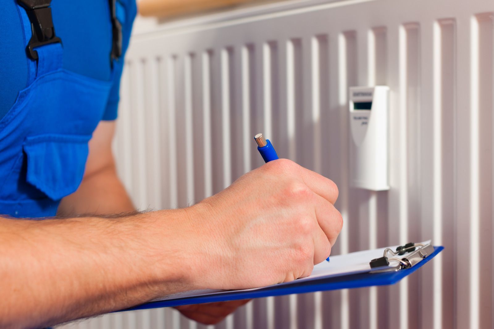 Person in blue overalls writing on a clipboard by a radiator.