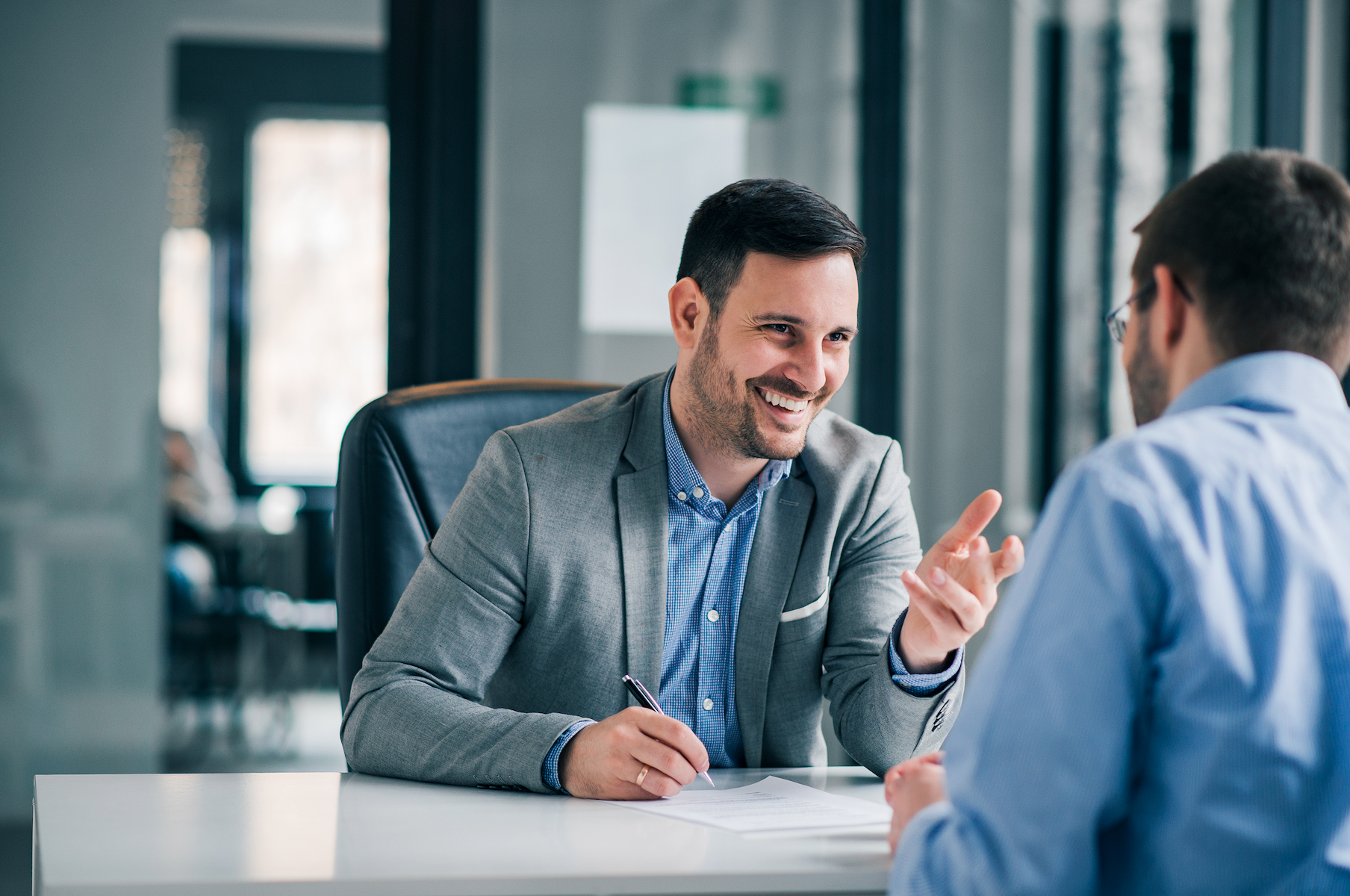 A man is sitting at a desk talking to another man.