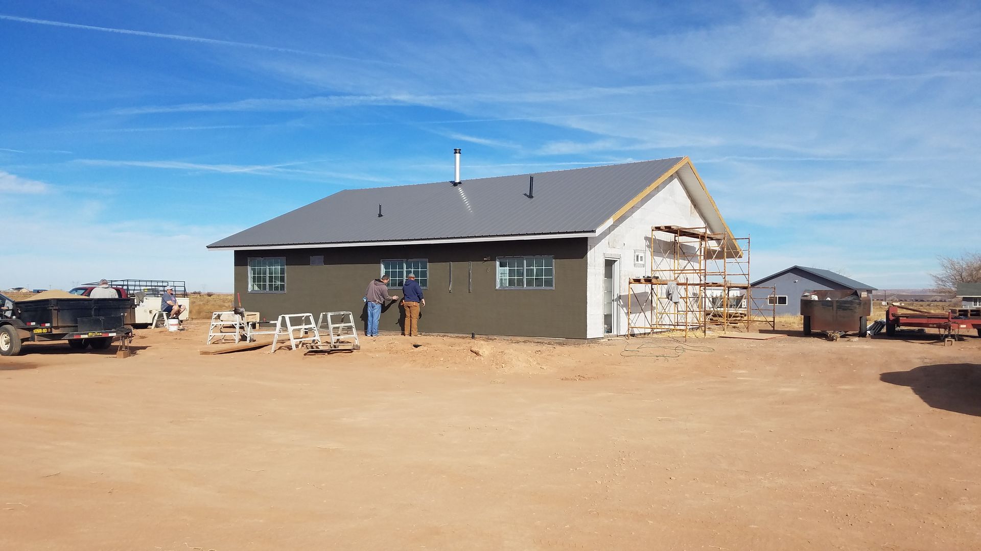 Two people work on the exterior wall of a partially finished, dark-sided house under a bright blue sky.